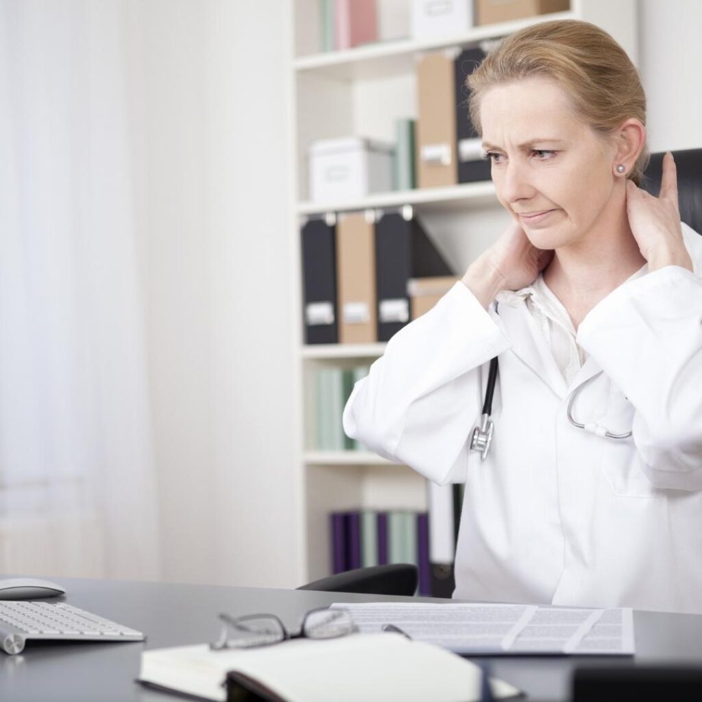 Tired Female Medical Doctor Sitting at her Office and Massaging the Back of her Neck While Looking Down.